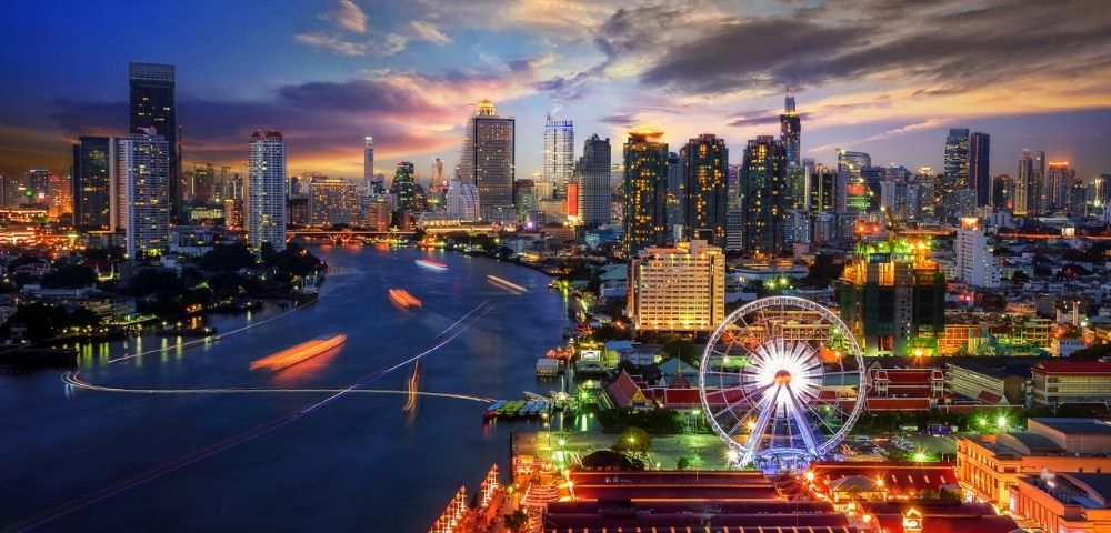 A vibrant city skyline at dusk with illuminated skyscrapers and a glowing Ferris wheel by a river. The sky transitions from orange to deep blue.