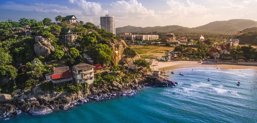 Aerial view of a coastal landscape with rocky cliffs, lush greenery, and houses, leading to a serene beach with turquoise waters and distant hills under a partly cloudy sky.