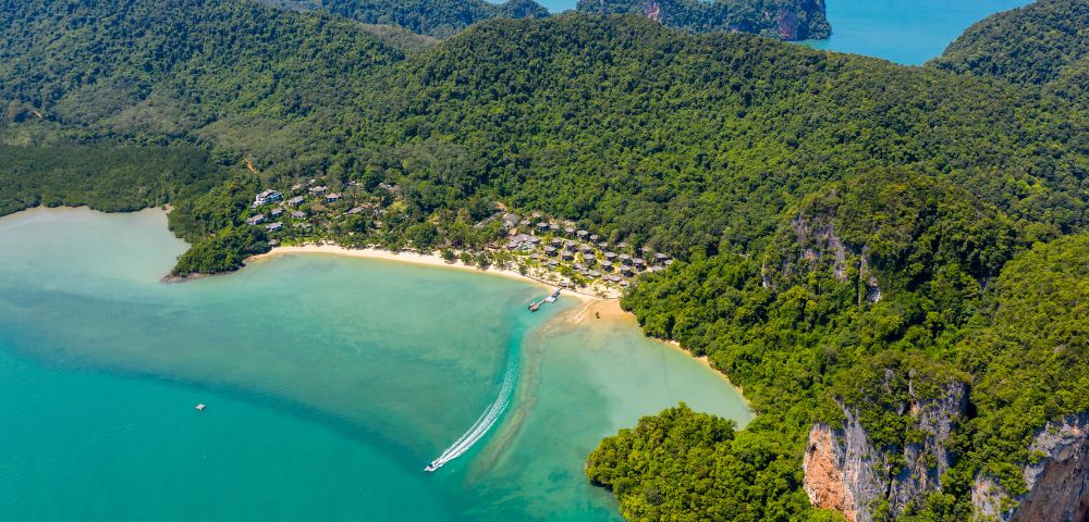 Aerial view of a tropical beach with turquoise water and dense green forest. A speedboat leaves a white trail, creating a serene, inviting atmosphere.