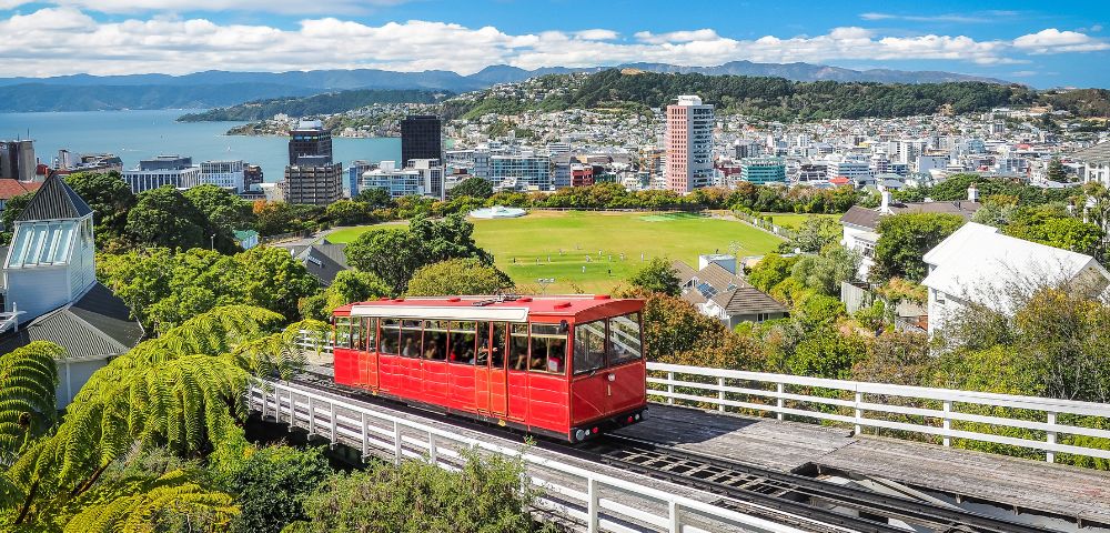 A red cable car ascends a track through lush greenery with a cityscape and ocean bay in the background on a sunny day, conveying a scenic and cheerful atmosphere.