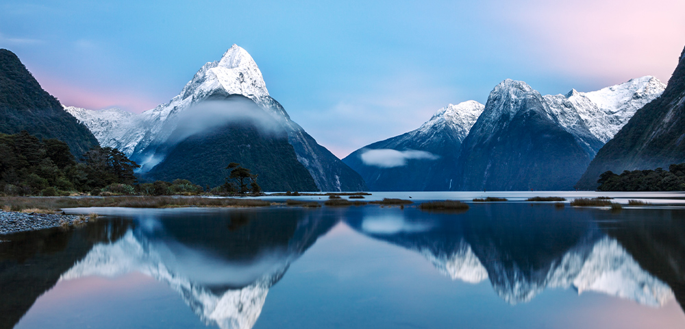 Snow-capped mountains reflected in a serene lake, surrounded by lush greenery. The sky has a soft pink hue, creating a tranquil and majestic scene.