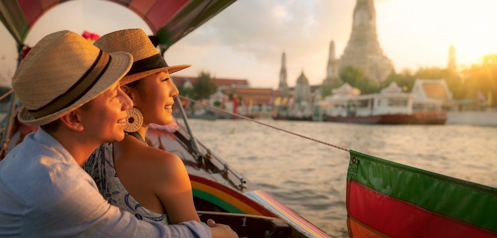 A couple in straw hats, smiling and embracing, enjoys a boat ride on a river at sunset, with a temple in the background. The scene conveys warmth and romance.
