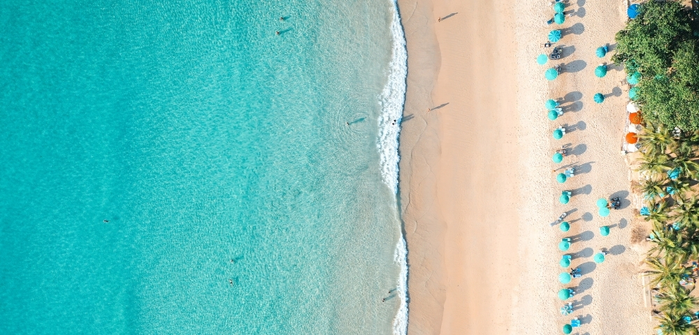 Aerial view of a beach with turquoise water and gentle waves. People swim near the shore, while others relax under umbrellas on the sandy beach.