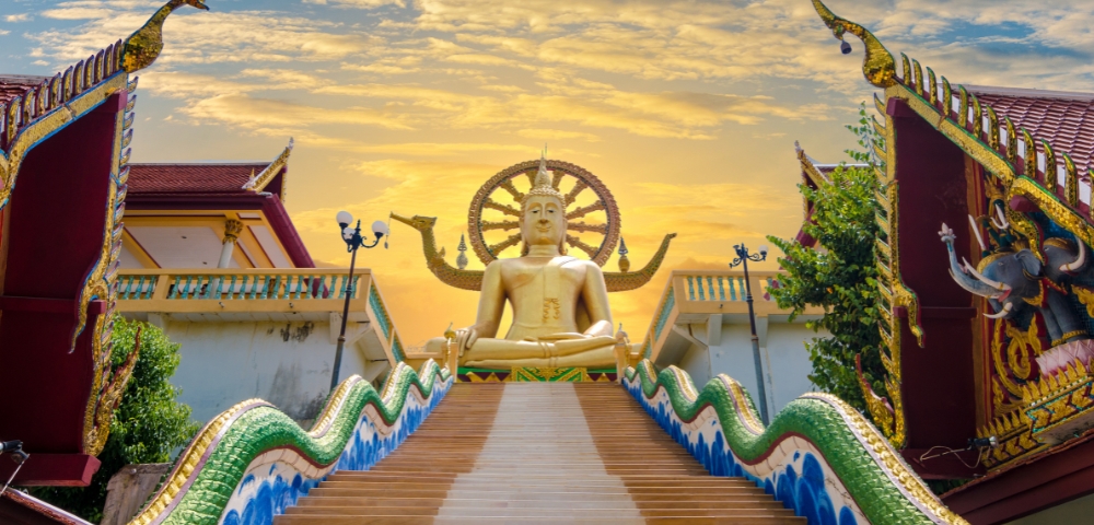 Golden Buddha statue on a temple platform with ornate, multicolored dragon railings. Vibrant sunset sky adds a serene and majestic atmosphere.