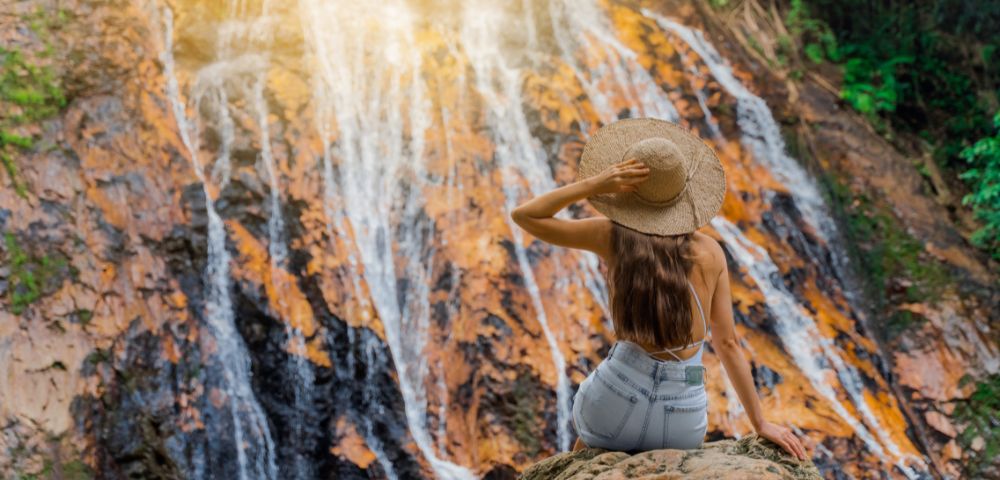 A woman sits on a rock facing a cascading waterfall, wearing a straw hat and blue jeans. Sunlight filters through the lush green foliage, creating a serene atmosphere.
