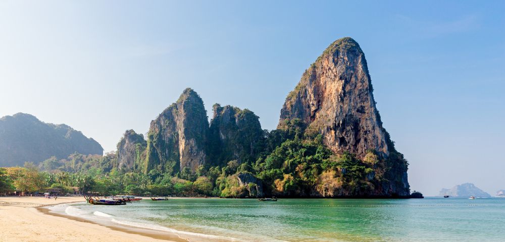 Serene beach scene with golden sand, calm turquoise water, and towering limestone cliffs covered in greenery under a clear blue sky.