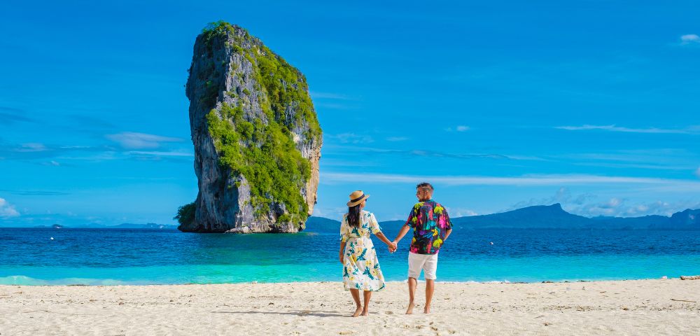 A couple holding hands on a sandy beach faces a towering limestone island under a clear blue sky. The scene conveys serenity and romance.