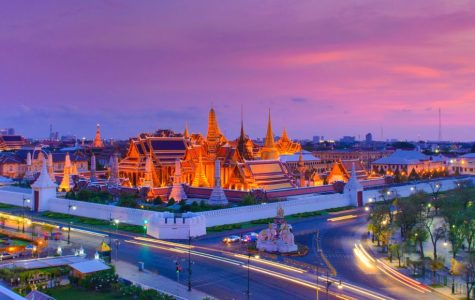 A vivid evening skyline of Bangkok featuring the illuminated Grand Palace with golden spires. The sky is a gradient of pink and purple, creating a serene ambiance.