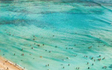 Aerial view of a vibrant beach with turquoise waters and numerous people swimming. A sailboat with blue stripes is near the shore, evoking a lively, tropical vibe.