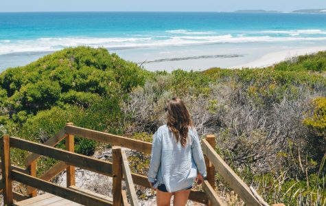 A woman walks down a wooden path toward a pristine beach with turquoise waves. Lush green bushes line the path, conveying a serene, inviting atmosphere.