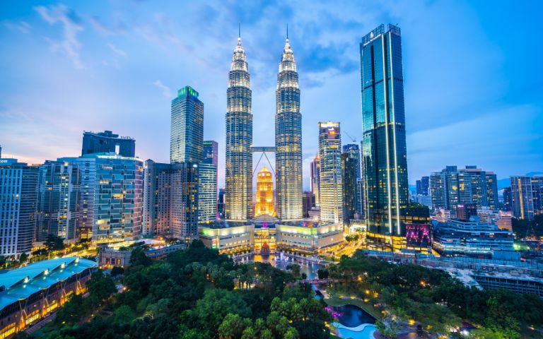 Kuala Lumpur skyline at dusk, featuring the illuminated Petronas Twin Towers surrounded by modern skyscrapers and a foreground of lush green parkland.