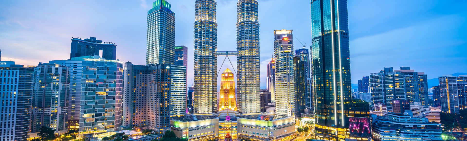 Kuala Lumpur skyline at dusk, featuring the illuminated Petronas Twin Towers surrounded by modern skyscrapers and a foreground of lush green parkland.