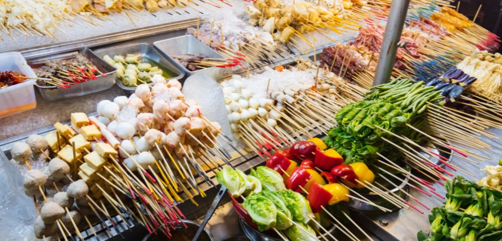 A vibrant street food stall displays an array of skewered ingredients, including vegetables, meat, and seafood. The colorful assortment creates a lively, appetizing scene.