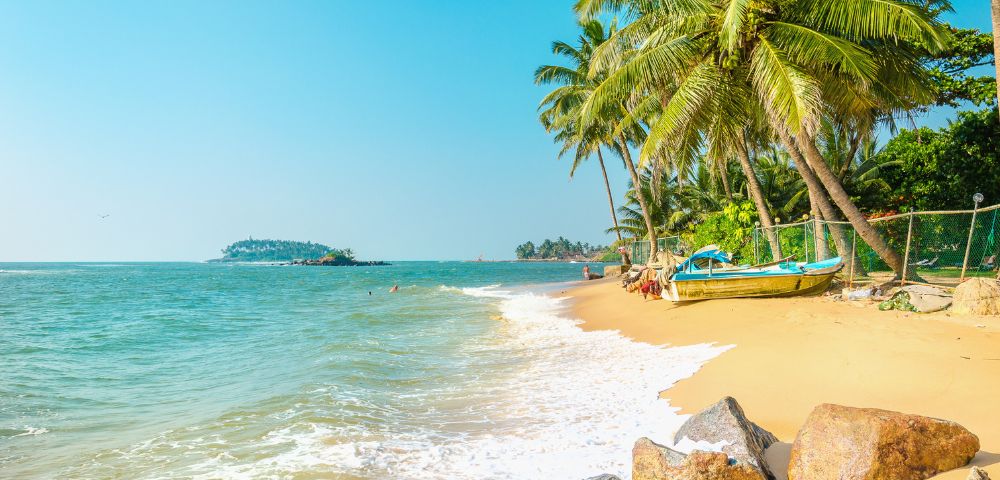 Tropical beach scene with golden sand, gentle waves, and lush palm trees. A colorful boat rests onshore, evoking a serene and sunny atmosphere.