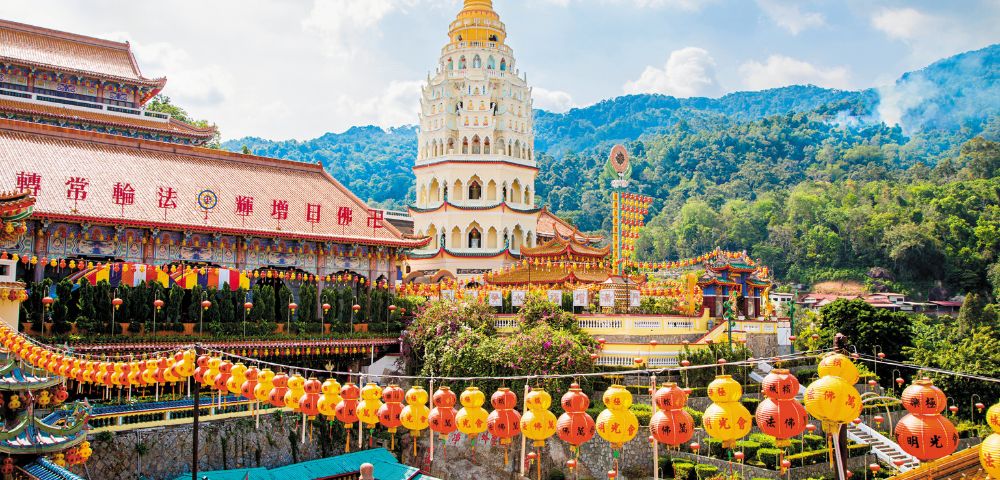 Vibrant temple complex with a multi-tiered pagoda surrounded by lush green hills. Colorful lanterns hang above, creating a festive atmosphere.