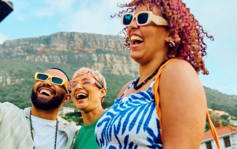 Three people wearing sunglasses laugh joyfully outdoors. A scenic mountain and palm trees are in the background, evoking a carefree, sunny vibe.