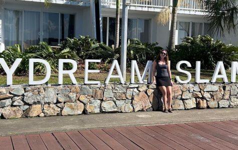 A person stands by a stone wall with large white letters spelling "Daydream Island" in front of a resort building with balconies and palm trees.