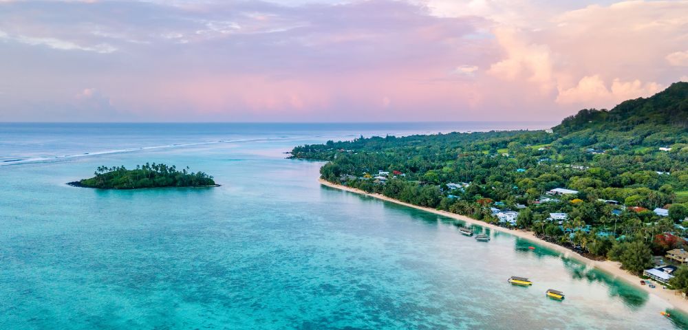 A serene coastal scene at sunrise with turquoise waters, a small island, and lush greenery along the shore under a pastel pink sky, conveying tranquility.