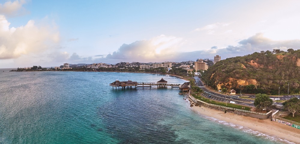Coastal cityscape featuring a serene beach, turquoise waters, and wooden piers. A road winds past lush hills, with city buildings under a cloudy sky.