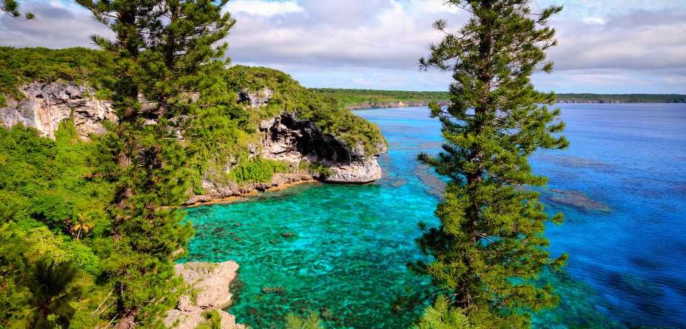A turquoise bay with clear water and rocky cliffs, surrounded by lush green forests and tall trees under a partly cloudy sky, evokes tranquility.