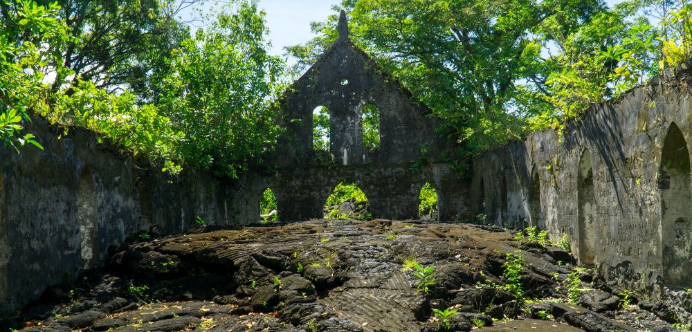 Overgrown ruins of a stone church with an open roof, surrounded by lush greenery. Sunlight filters through trees, creating a serene, timeless atmosphere.