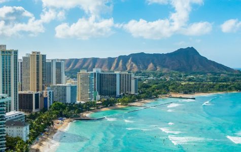Panoramic view of Honolulu beachfront with high-rise buildings, turquoise ocean, sandy beach, and Diamond Head volcanic crater in background.