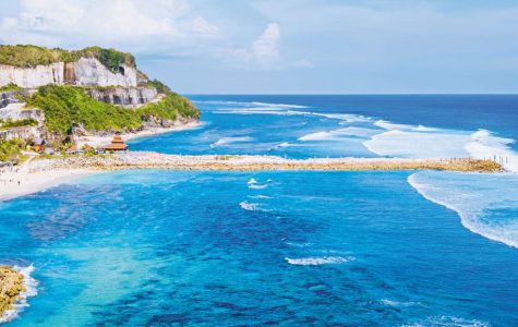 Aerial view of a vibrant blue ocean meeting a sandy beach, with rugged cliffs on the left. The sky is clear, with a few fluffy clouds, conveying tranquility.