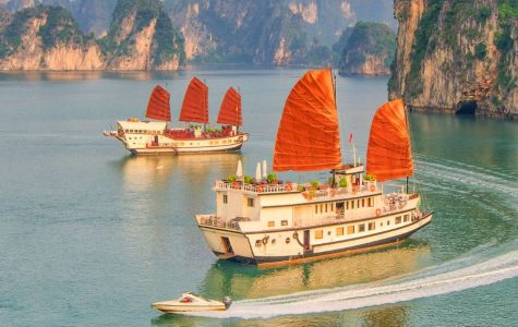 Traditional wooden boats with bright red sails glide across a calm turquoise bay beneath towering limestone cliffs, creating a serene, scenic mood.