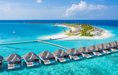 Aerial view of a tropical island resort with overwater bungalows and villas on pristine white sand beaches, surrounded by vibrant turquoise waters under a bright blue sky.