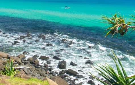 Panoramic view of a rocky coastline with lush green vegetation and vibrant turquoise ocean water, featuring waves crashing against the shore and a small boat in the distance.