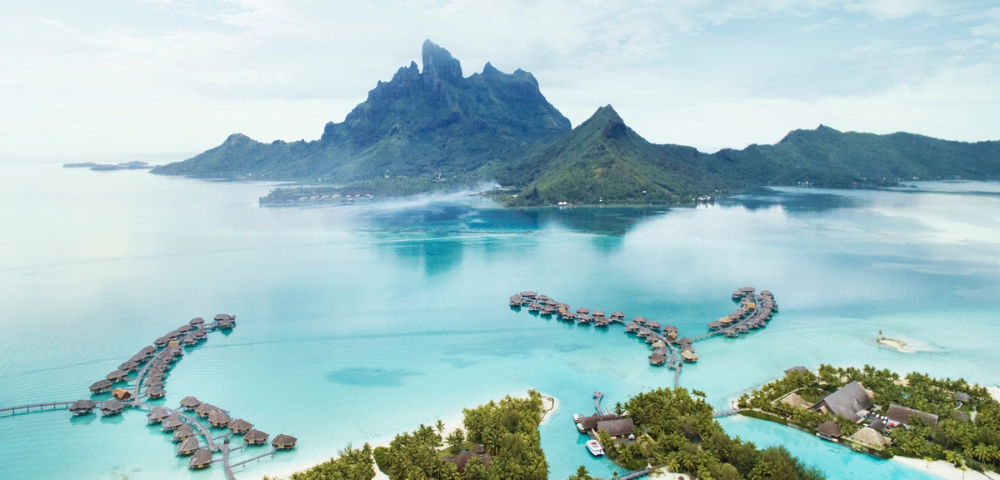 Aerial view of overwater bungalows extending into a turquoise lagoon with a large, dramatic volcanic mountain rising in the background under sky.