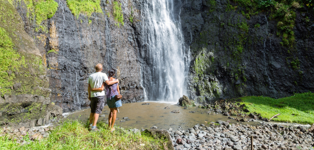 Two hikers stand beside a rocky pool beneath a tall waterfall cascading down a moss-covered cliff.
