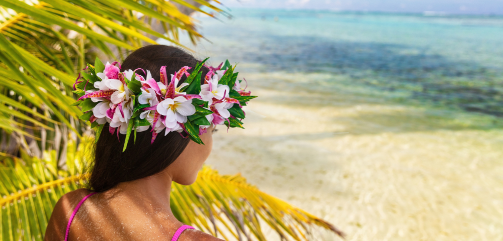A person wearing a colorful floral crown looks out toward a clear turquoise lagoon, framed by arching palm fronds in the foreground. The tropical shoreline, bright greenery, and calm ocean water create a peaceful island setting under soft daylight.