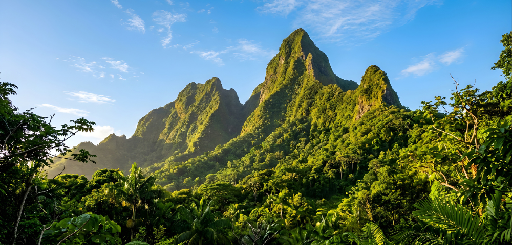 A dramatic series of steep, green mountain peaks rises above a dense tropical rainforest, illuminated by warm sunlight beneath a bright blue sky with scattered white clouds. Lush tropical vegetation fills the foreground, creating a vibrant landscape of rich greens and rugged volcanic-looking ridges.