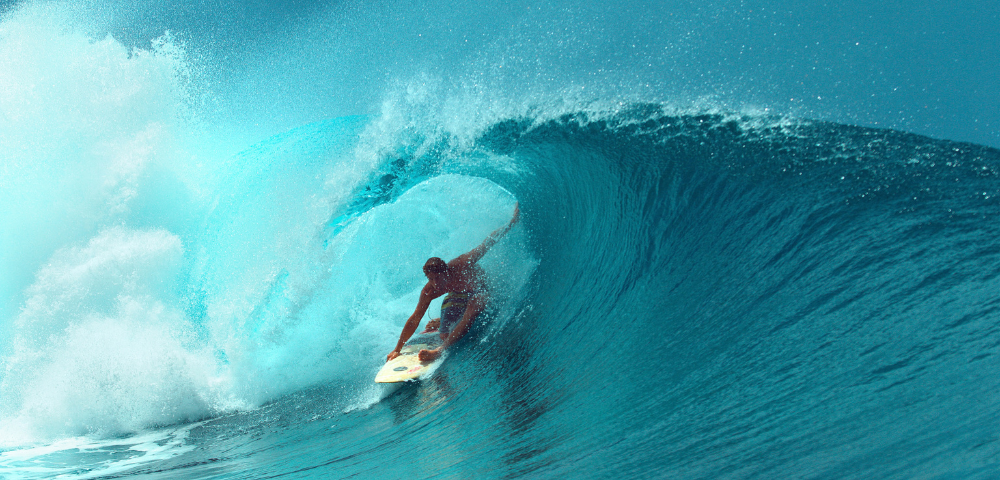 A surfer rides a white surfboard deep inside the barrel of a large turquoise wave, surrounded by crashing spray and foaming water. The powerful ocean wave curves overhead, creating a dramatic tunnel of water against a bright, sunlit seascape.